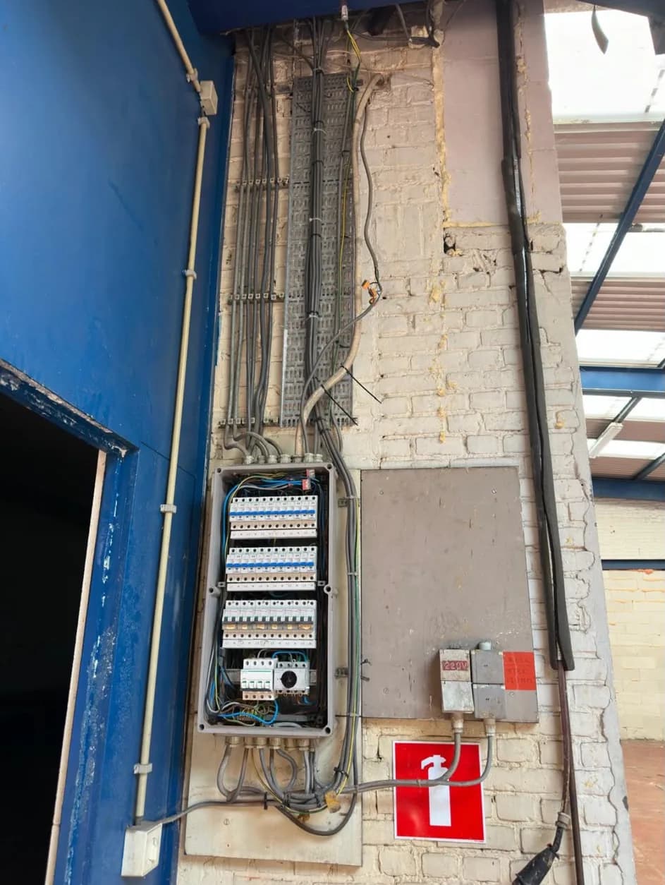 Oude verdeelkast zonder deksel tegen een bakstenen muur in een bedrijfshal, met losse kabels naar boven